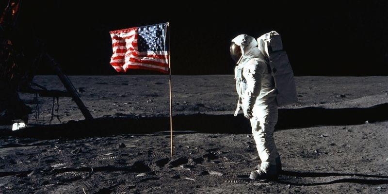 Astronaut looking towards American Flag Standing at the surface of Moon